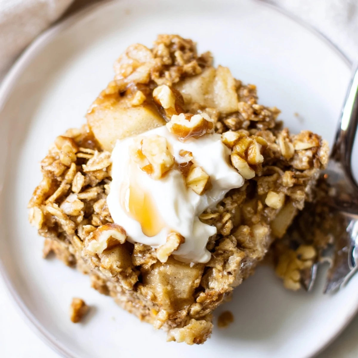 Golden squares of Baked Apple Oatmeal With Greek Yogurt topped with a creamy dollop and toasted pecans on a rustic plate.  