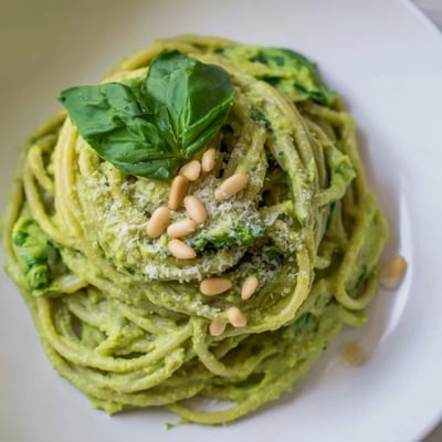 A close-up of creamy avocado and spinach pasta with fresh basil and pine nuts garnishing a warm bowl.  